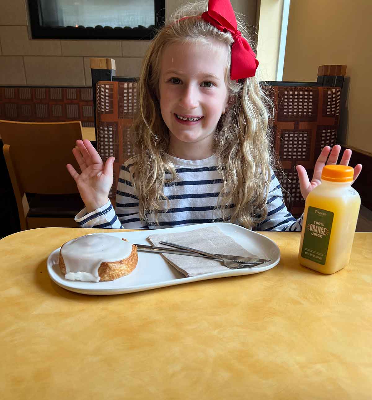 Little girl with a red bow in her hair sitting at a restaurant with a cinnamon roll and orange juice.